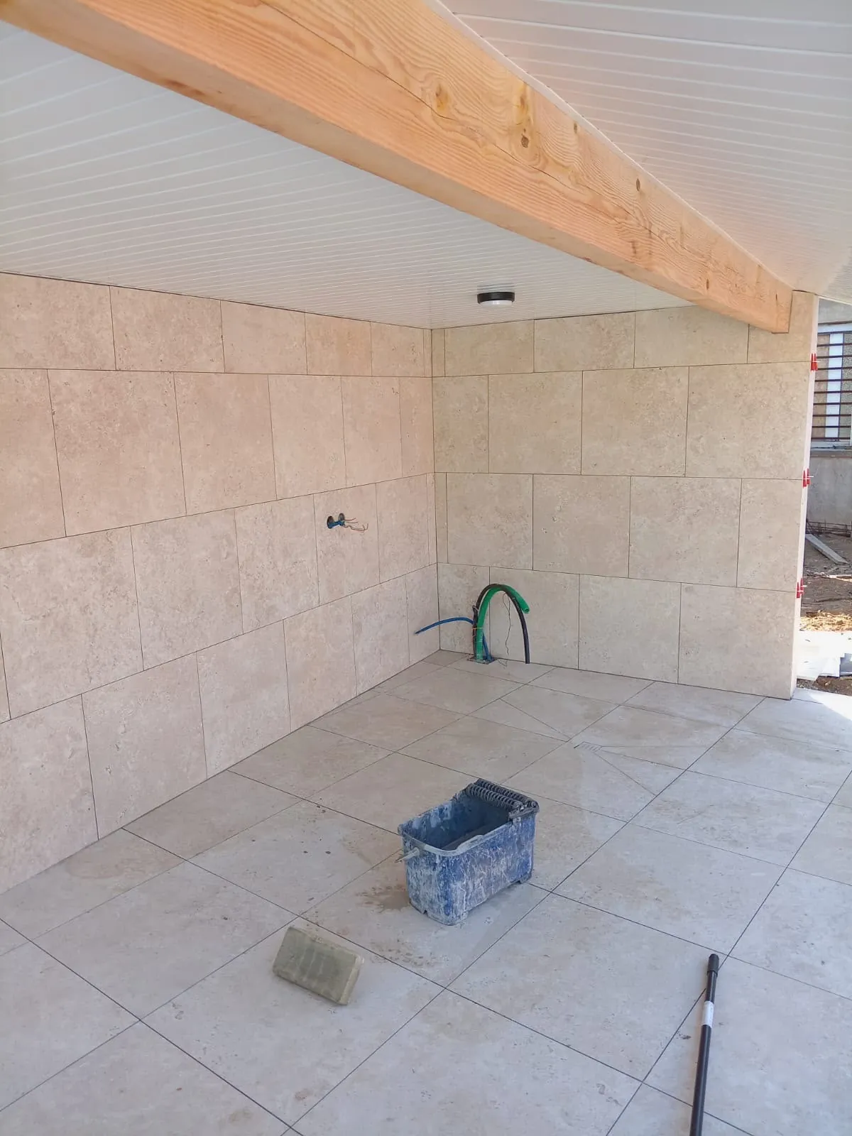 Abri extérieur sous une poutre en bois et un plafond en lames blanches, murs et sol en grands carreaux beige façon pierre, attentes d’eau au pied du mur
