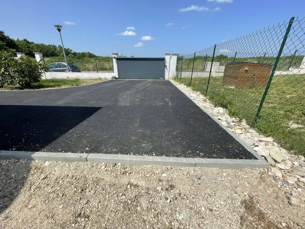 Allée d’accès en enrobé noir tenue par des bordures en béton, portail au fond, grillage et pelouse sur le côté