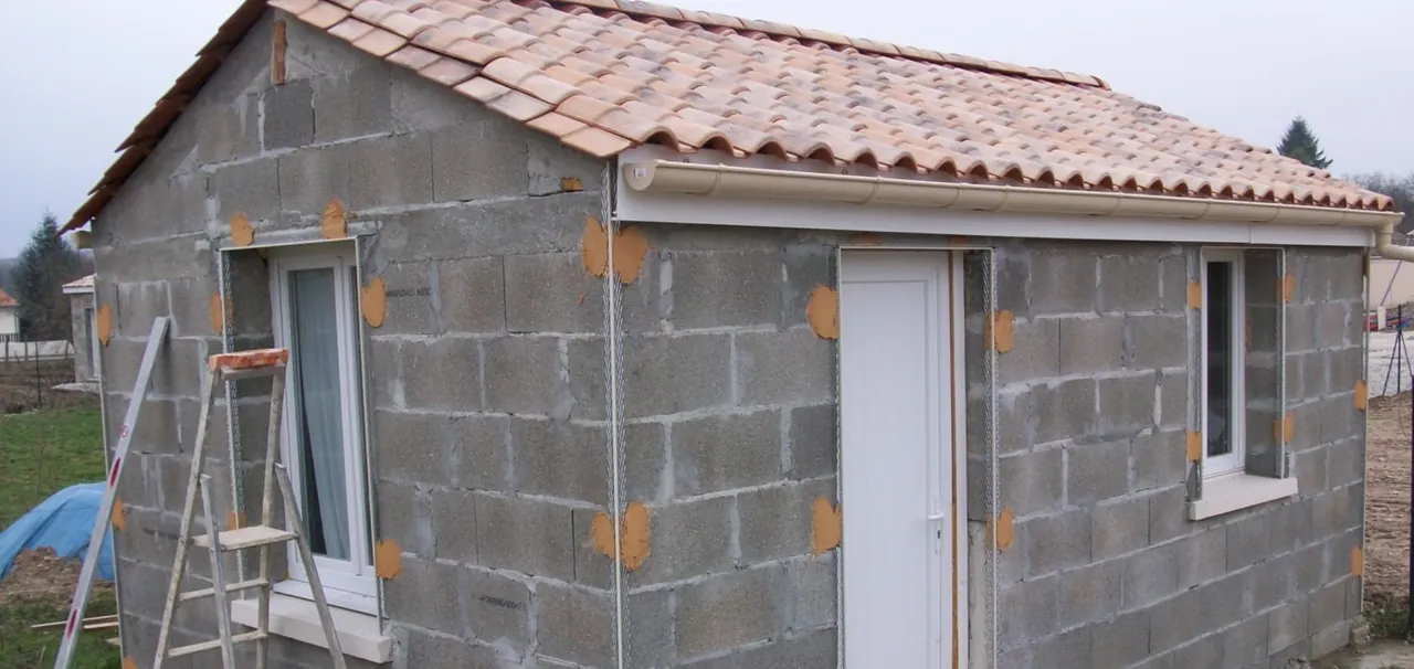 Petit bâtiment en blocs béton couvert de tuiles, gouttière posée, porte et fenêtres en place, échelle contre le mur