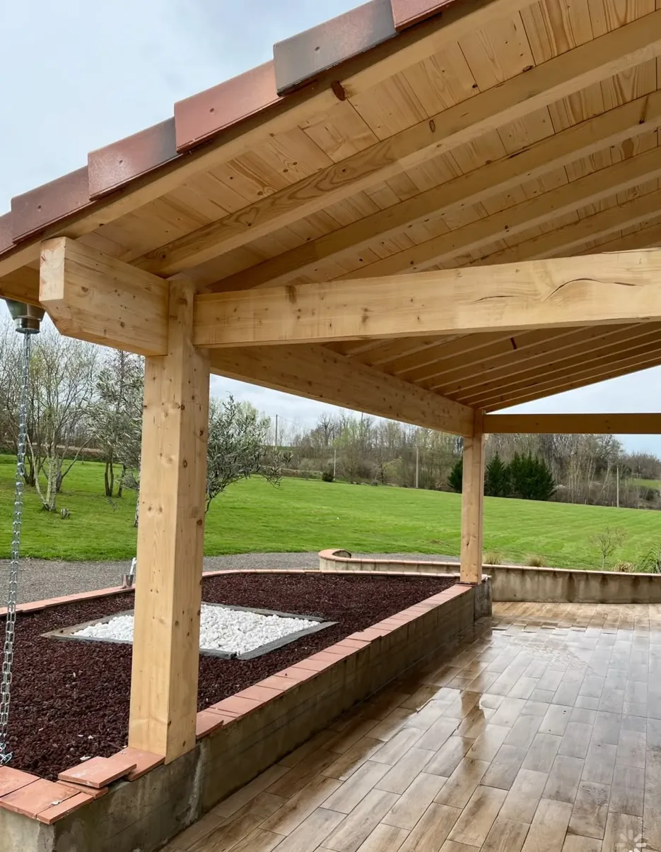 Auvent à poteaux et poutres en bois clair couvert de tuiles, au-dessus d’une terrasse en carrelage façon bois, jardinières maçonnées