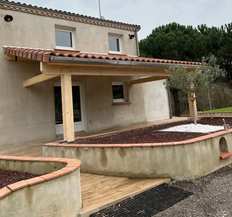 Auvent en bois couvert de tuiles contre la façade d’une maison, terrasse en carrelage façon bois et jardinière maçonnée au premier plan
