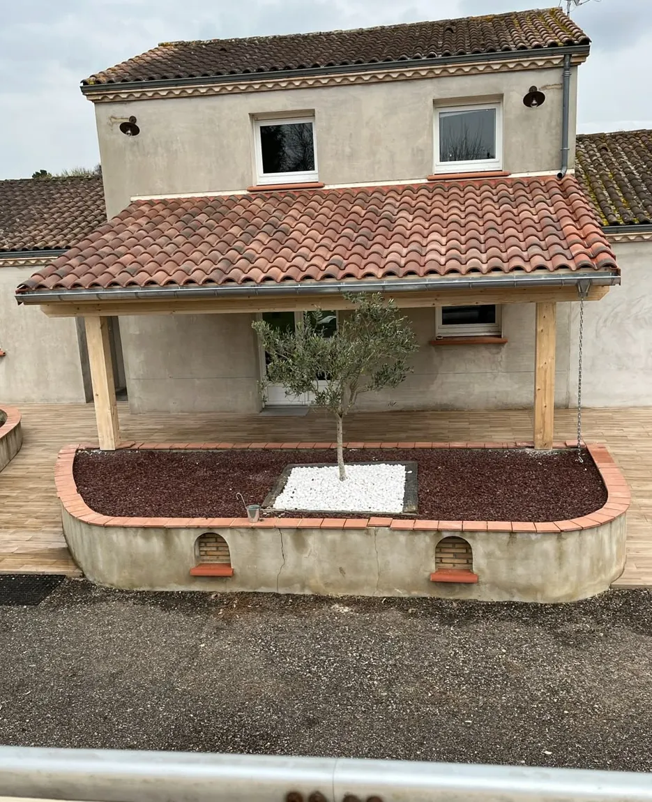 Façade de maison avec un auvent en bois couvert de tuiles, terrasse carrelée et jardinières maçonnées arrondies devant