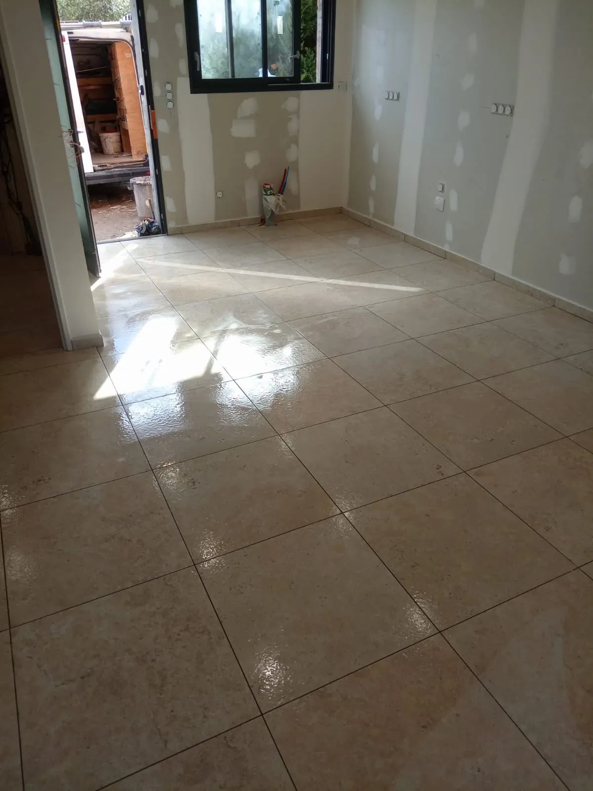 Pièce aux murs en plaques de plâtre jointées, sol en carreaux beige brillants, porte ouverte sur l’extérieur