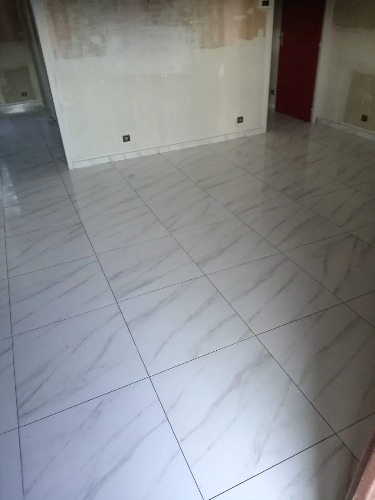 Pièce en rénovation : sol en grands carreaux blancs veinés de gris, murs en reprise, porte rouge au fond