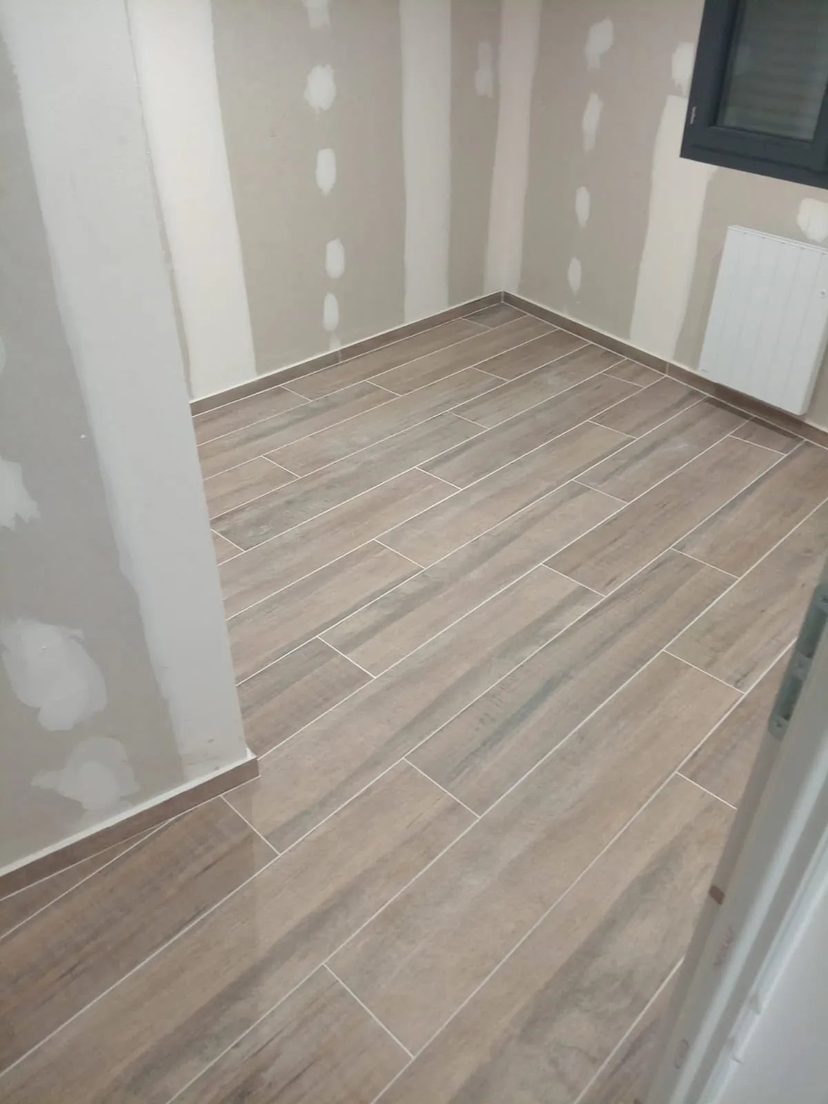 Chambre aux murs en plaques de plâtre jointées, sol en carrelage imitation parquet et plinthes assorties, radiateur sous la fenêtre