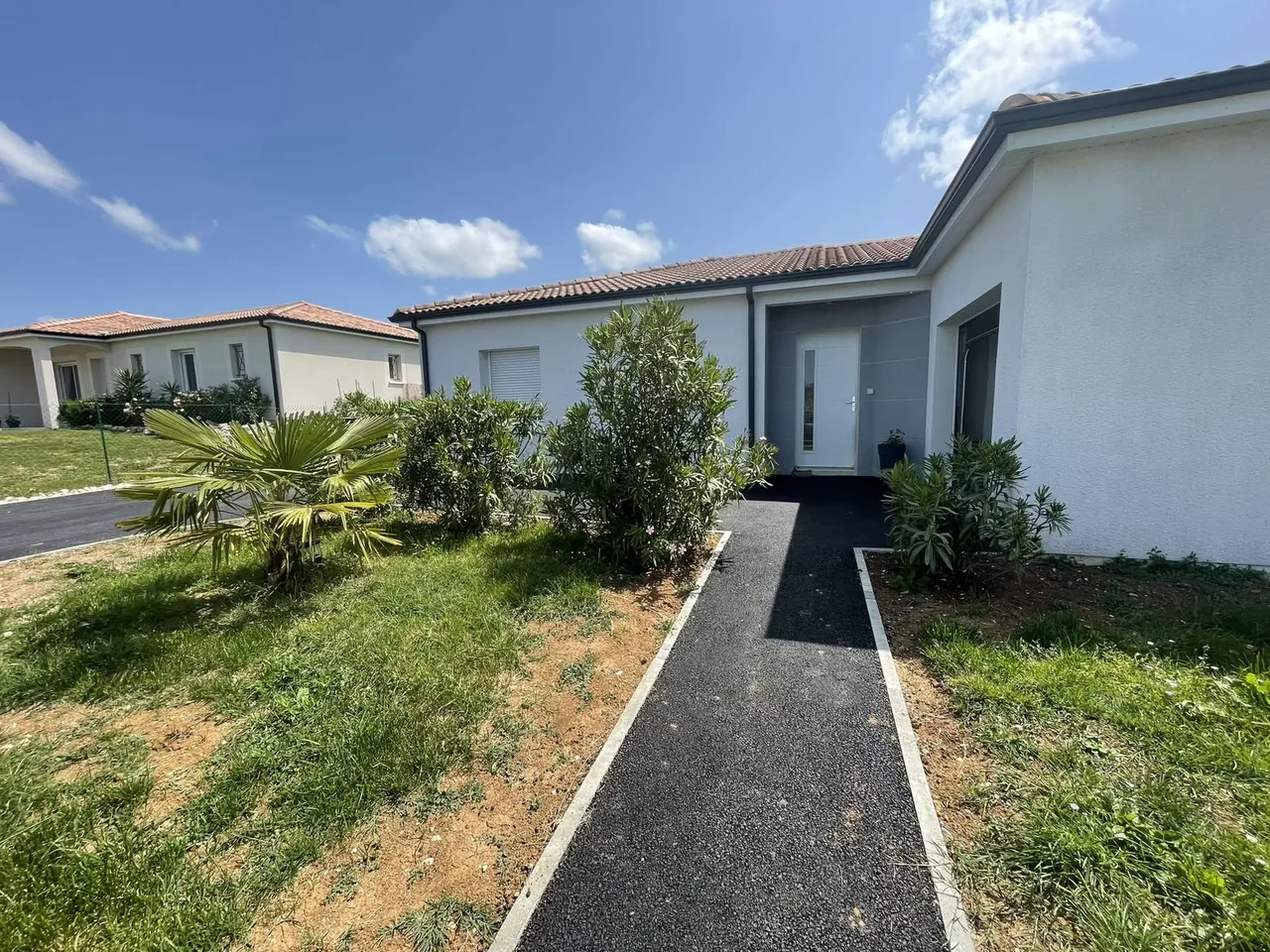 Chemin en enrobé noir bordé de bordures blanches qui mène à la porte d’entrée d’une maison, massifs plantés de part et d’autre