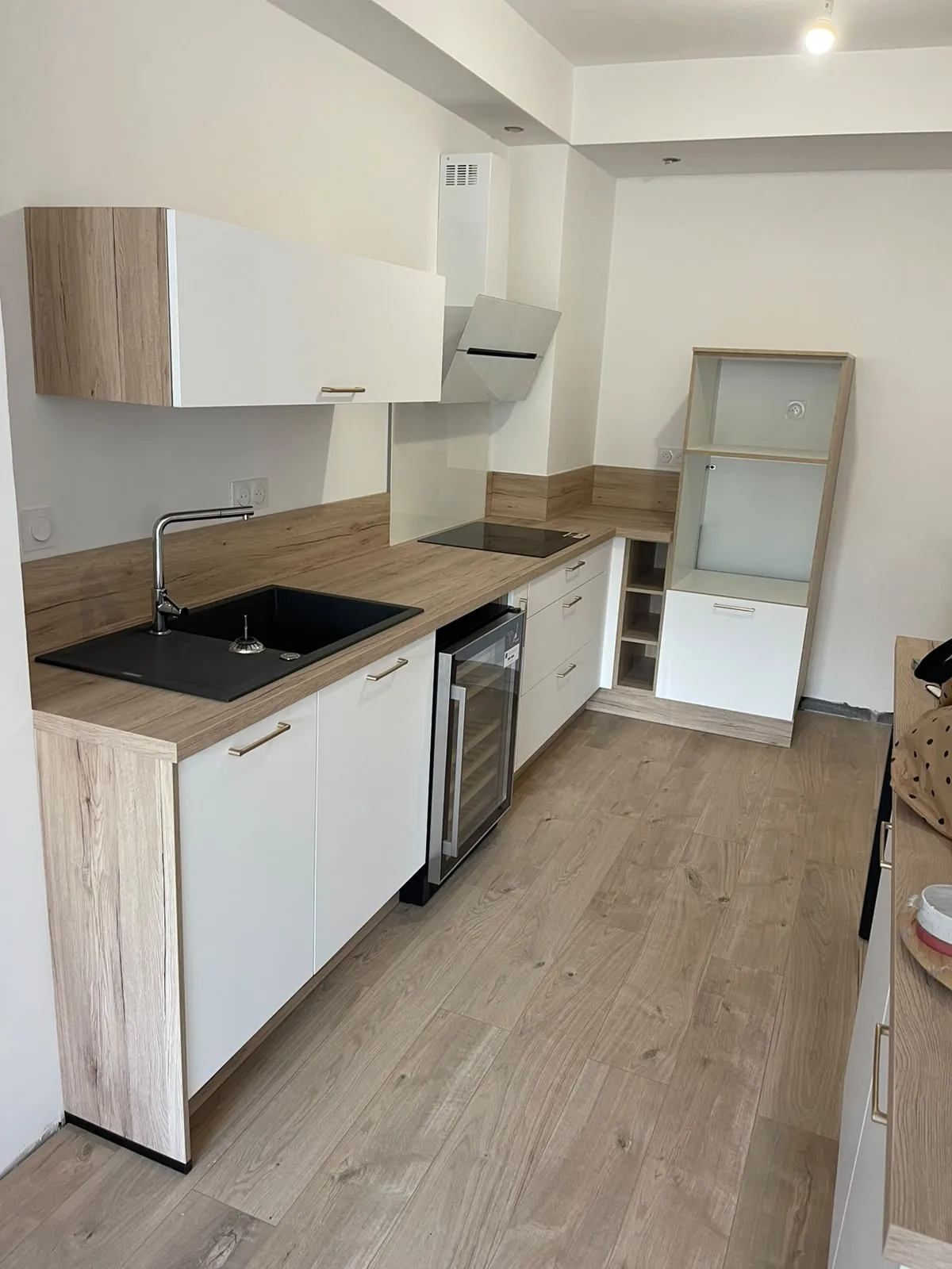 Cuisine en L aux façades blanches et au plan de travail façon bois, évier noir, plaque de cuisson et colonne pour le four, sol façon parquet