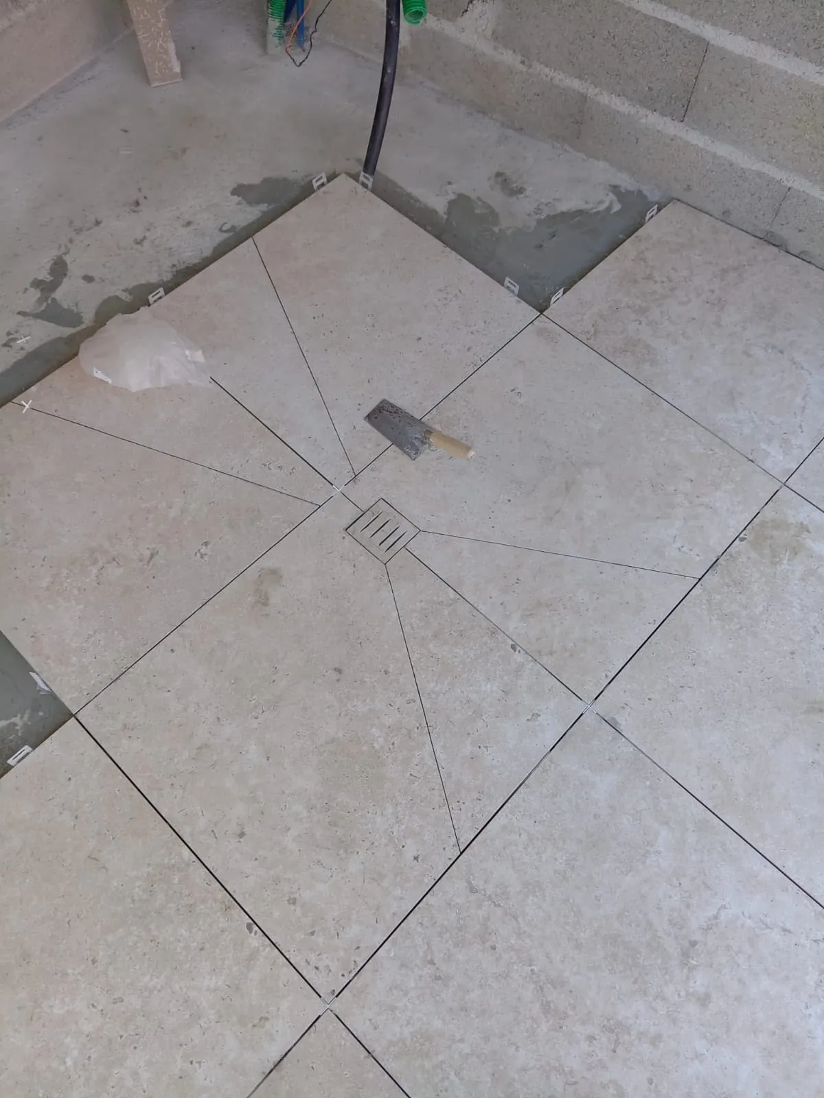 Sol de douche à l’italienne en grands carreaux beige, découpés en pointe vers la bonde carrée du centre, truelle posée sur le carrelage