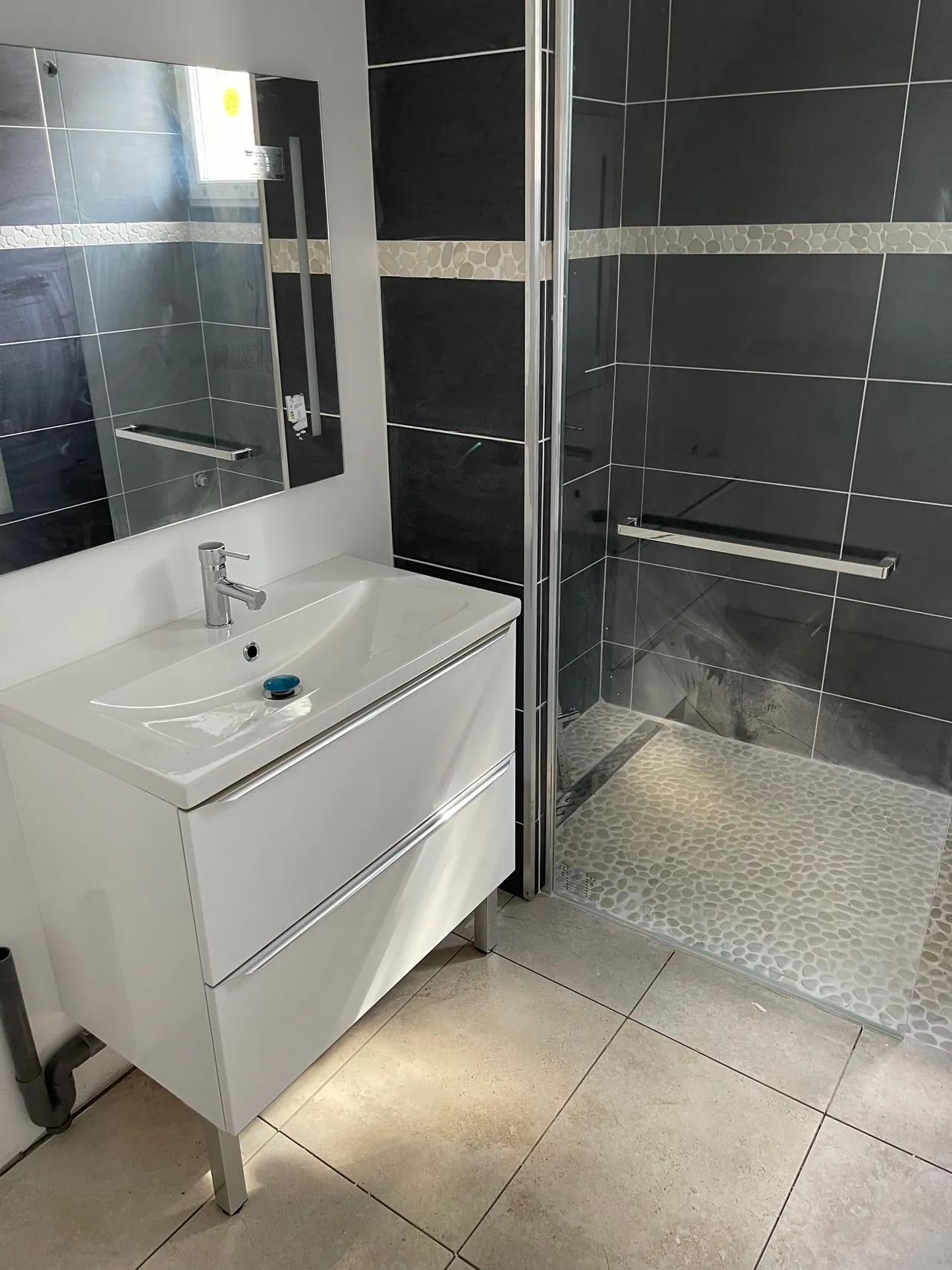 Salle d’eau terminée : meuble vasque blanc, miroir, douche à l’italienne au sol en galets, faïence anthracite