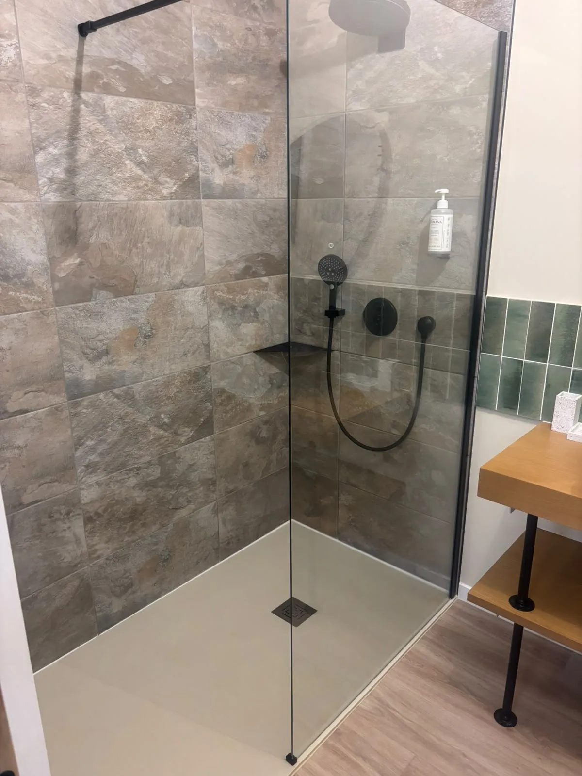 Douche aux murs en grands carreaux façon pierre, paroi vitrée à profil noir, colonne de douche et robinetterie noires, receveur blanc