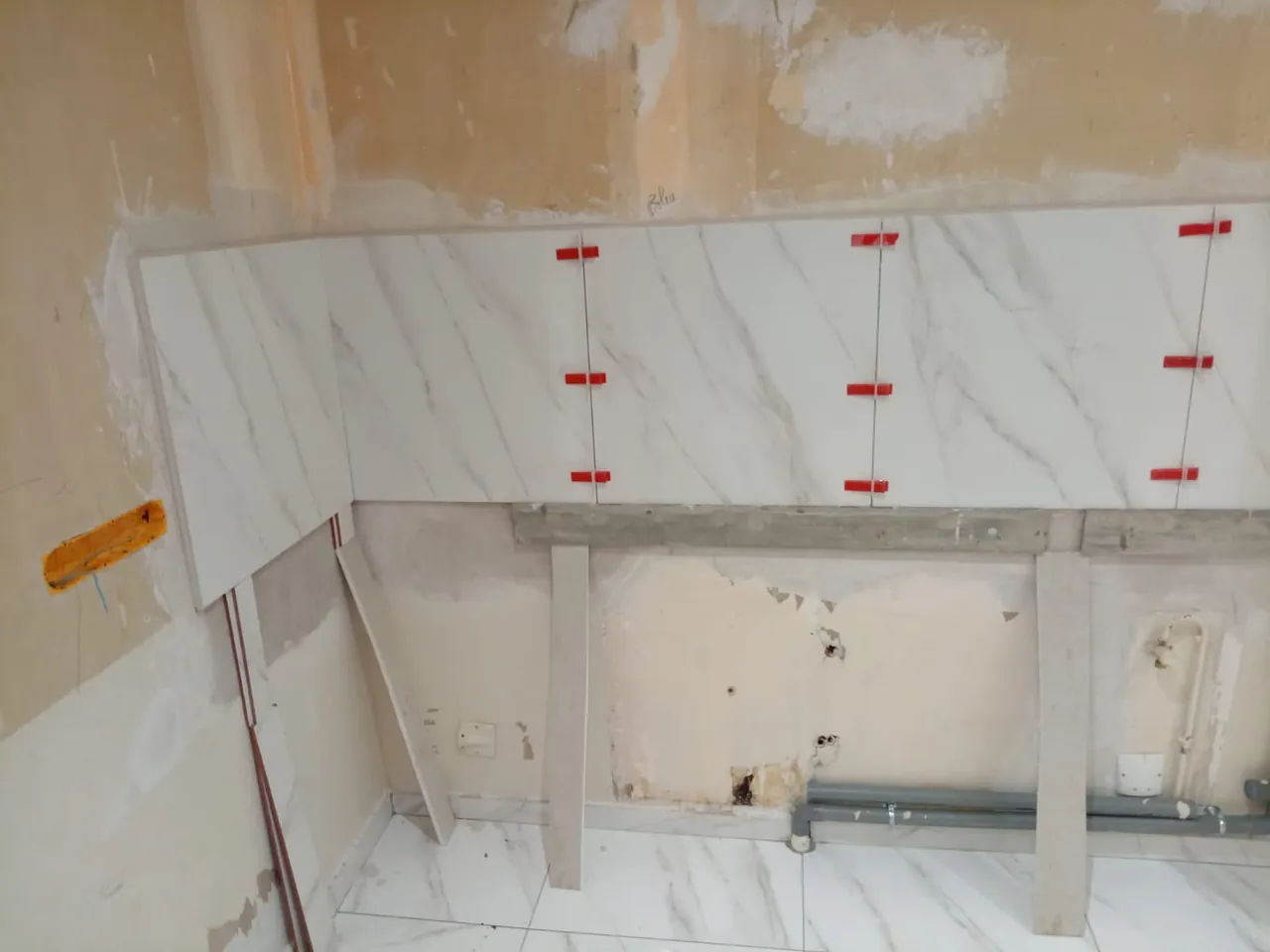 Mur de cuisine en rénovation : grands carreaux blancs veinés posés au-dessus d’un tasseau de maintien, cales rouges entre les carreaux, évacuations en attente au pied du mur