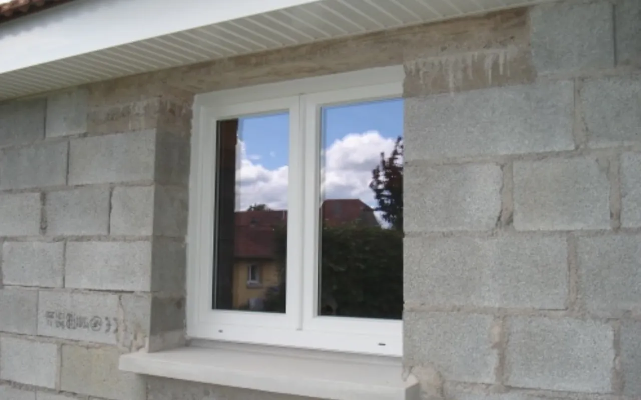 Fenêtre blanche posée dans un mur en blocs béton, appui en béton, avancée de toit à lames blanches au-dessus