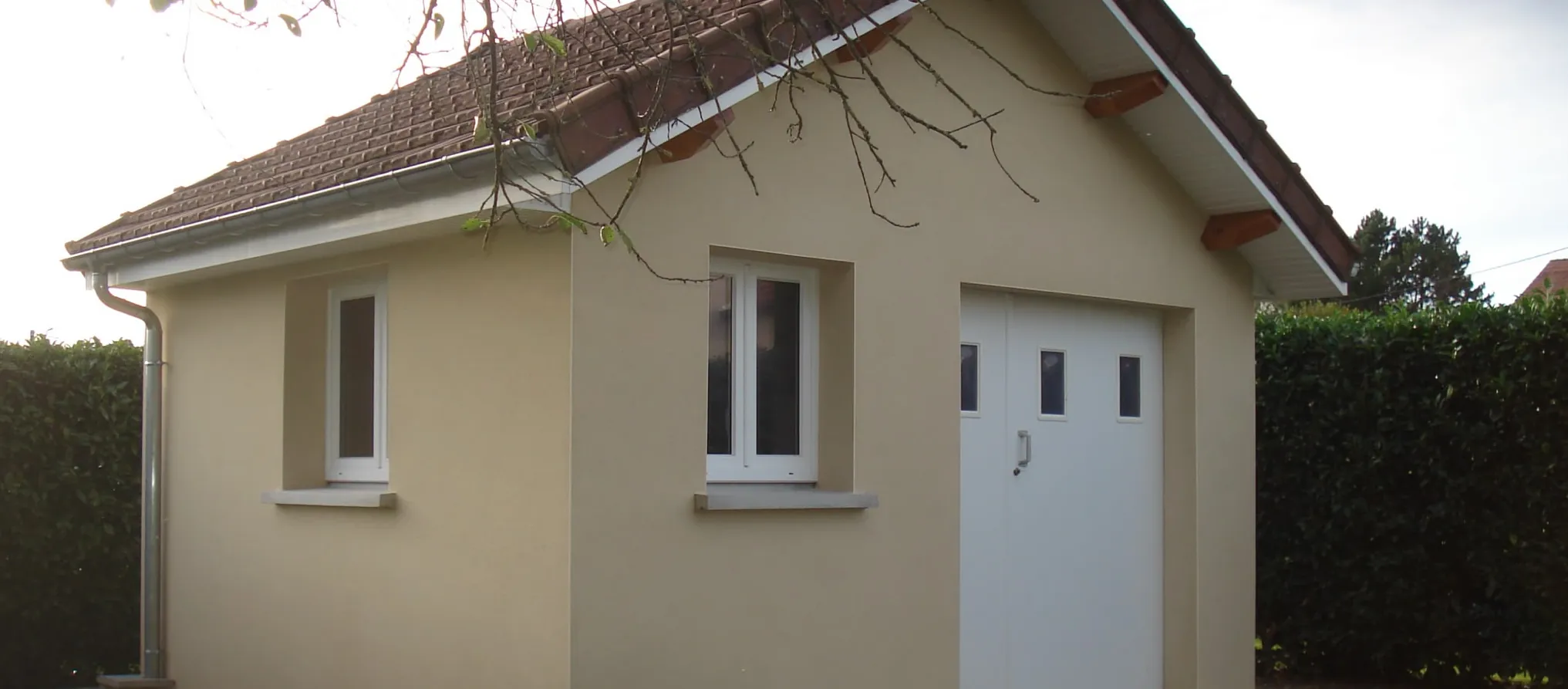 Garage aux murs enduits beige, toit en tuiles, fenêtres et porte de garage blanches, haie à côté