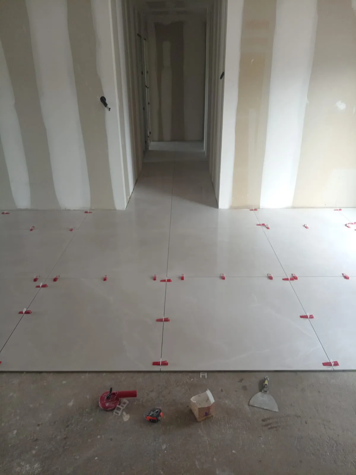 Couloir d’une maison en travaux : grands carreaux clairs et leurs cales de nivellement rouges, outils posés sur la chape au premier plan
