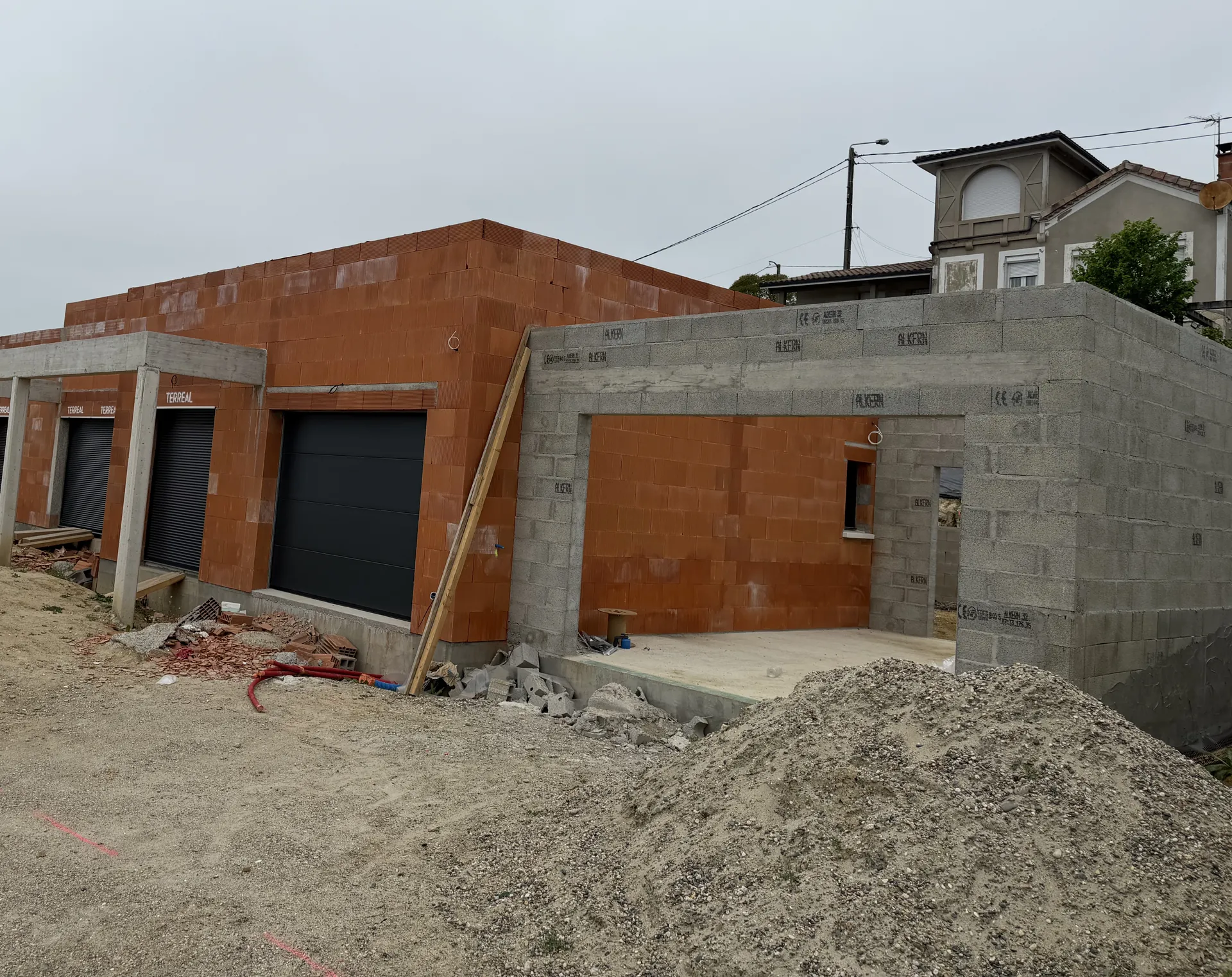 Grande maison neuve en briques avec son auvent sur poteaux et son garage accolé en blocs béton, tas de gravier du chantier devant