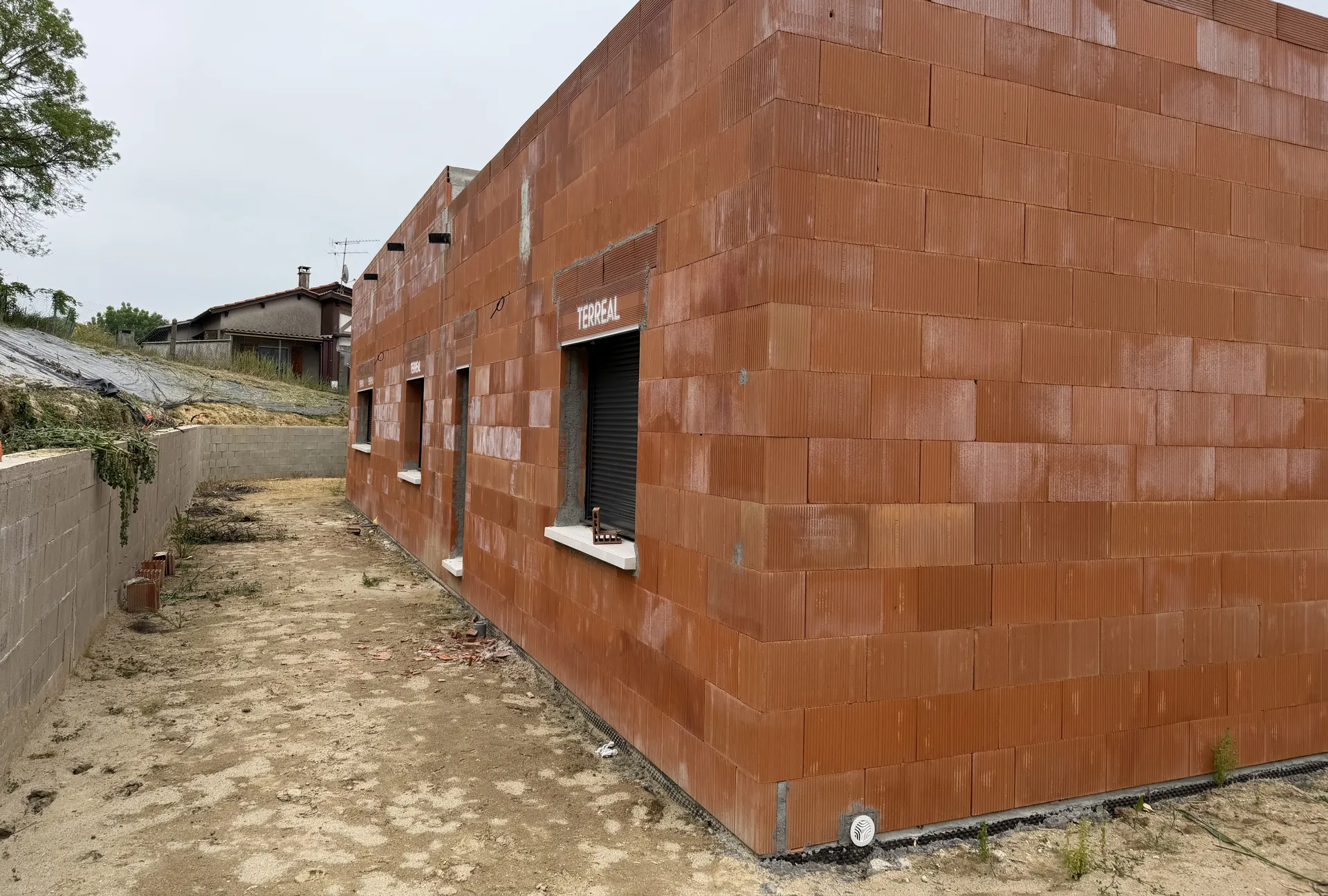 Angle d’une grande maison neuve de plain-pied : murs en briques de terre cuite montés, fenêtres à volets roulants posées, terre du chantier autour