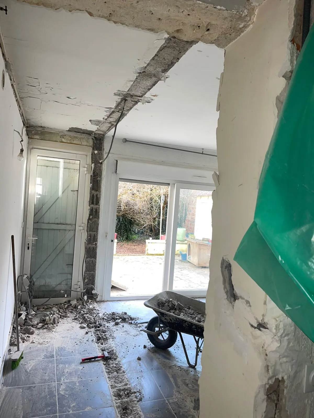 Pièce en rénovation : un mur vient d’être abattu, sa trace au sol et au plafond, gravats dans la brouette, baie vitrée ouverte sur la cour