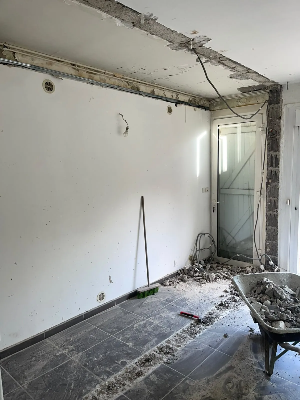 Pièce en rénovation après la démolition d’un mur : sa trace au sol et au plafond, brouette pleine de gravats, porte vitrée au fond