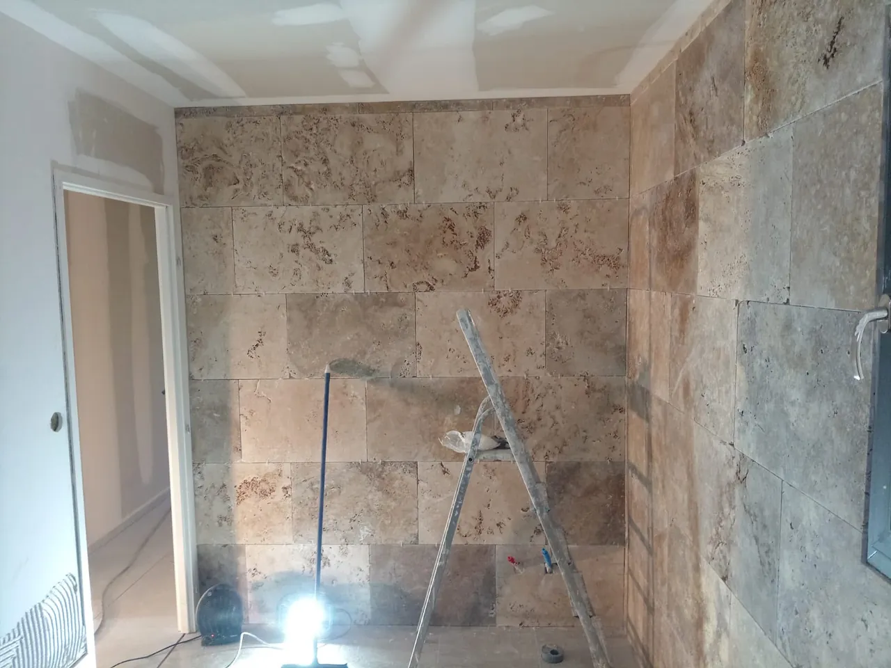 Pièce dont les murs sont habillés de grandes plaques de pierre beige et rouille, escabeau et projecteur de chantier au pied du mur