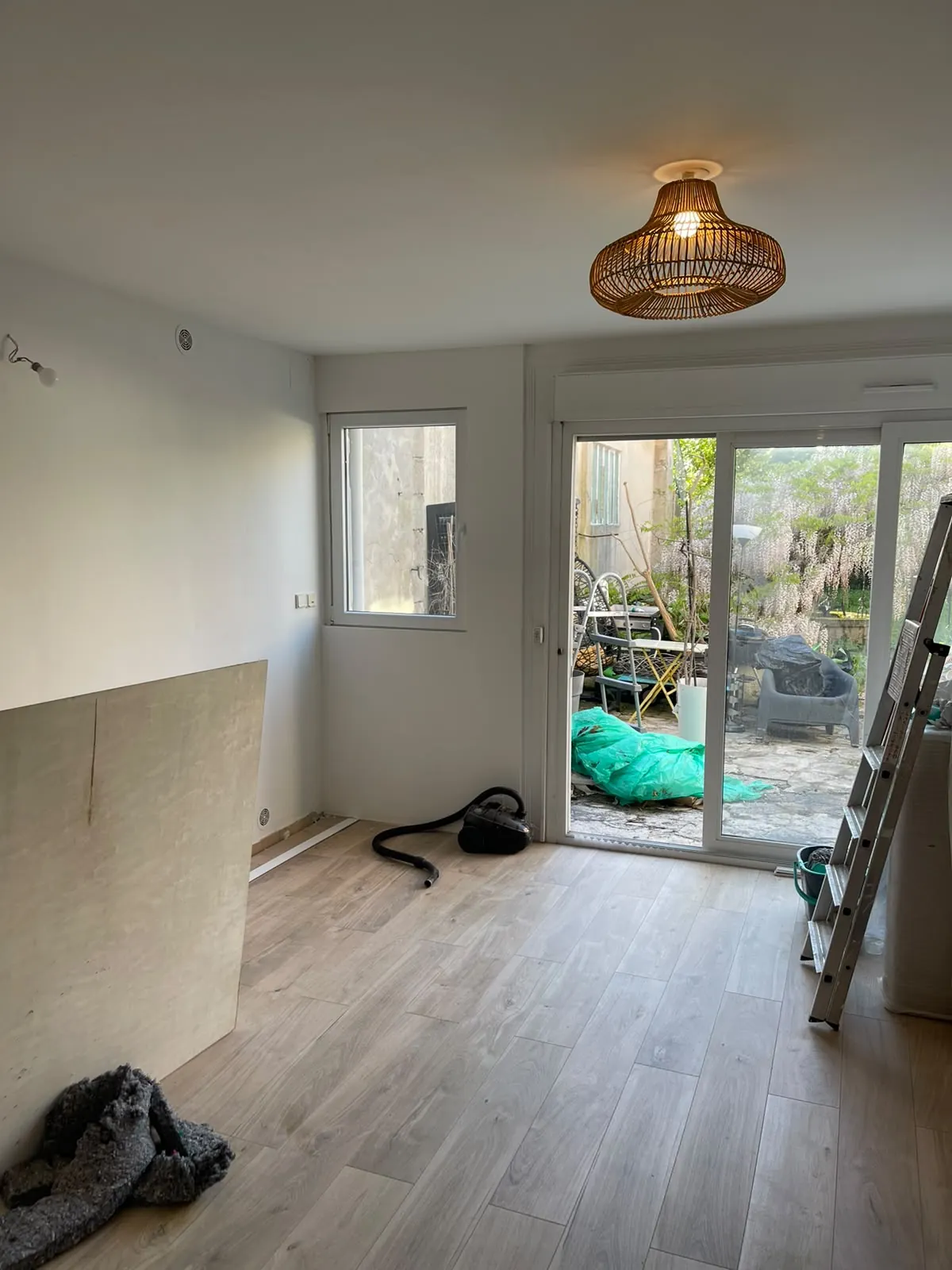 Pièce rénovée aux murs blancs, sol façon parquet posé, suspension en osier, baie vitrée sur la cour, échelle et aspirateur encore là