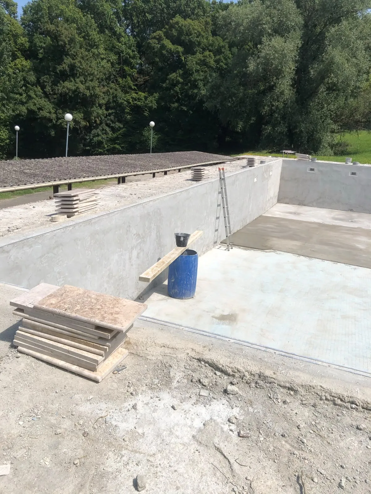 Bassin de piscine en béton en construction, pile de margelles en pierre au premier plan, échelle contre la paroi, arbres au fond