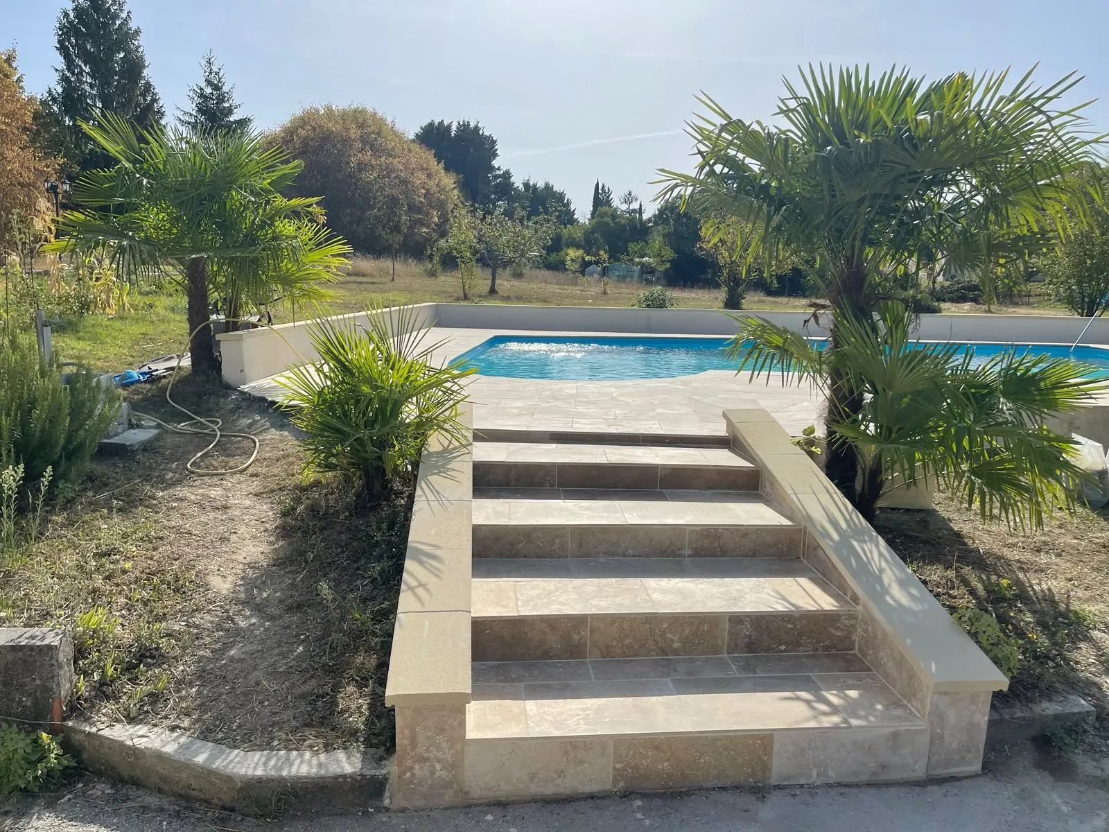 Escalier en dalles de pierre entre deux murets coiffés, encadré de palmiers, qui monte à la plage d’une piscine