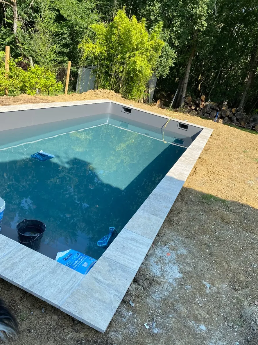 Petite piscine au liner bleu en cours de remplissage, margelles en pierre claire posées, terre remuée autour