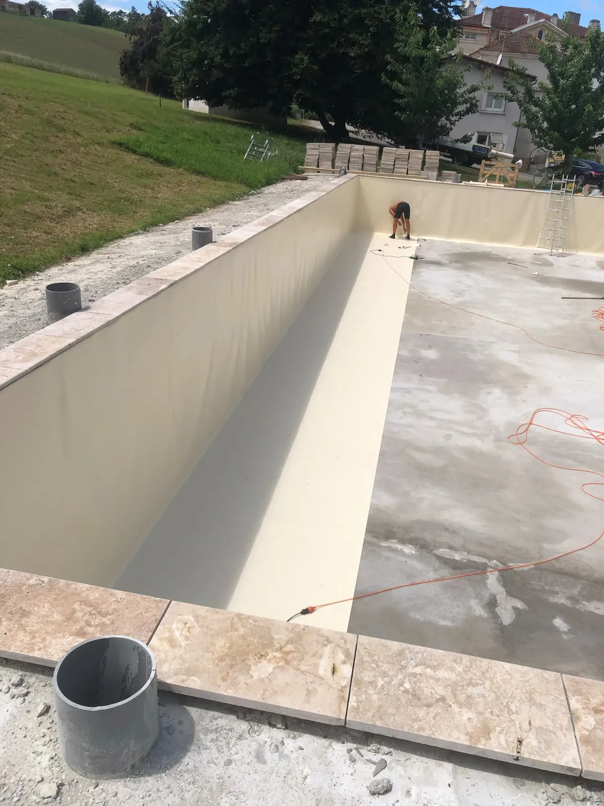 Long bassin de piscine au liner beige en cours de finition, margelles en pierre claire sur le bord, piles de dalles au fond