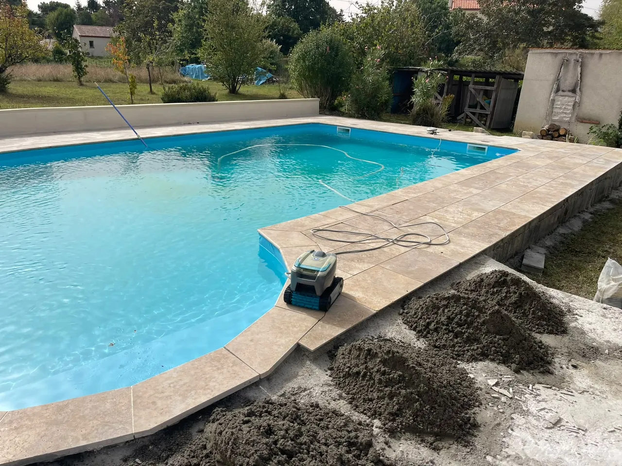 Piscine aux bords arrondis entourée de margelles en pierre claire, robot de nettoyage sur la plage, tas de mortier à côté