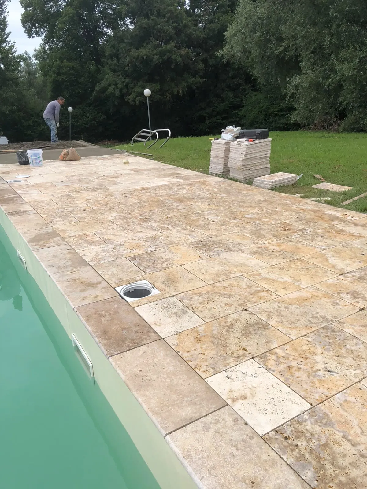 Plage de piscine en dalles de pierre beige de plusieurs formats, skimmer au bord du bassin, piles de dalles sur la pelouse
