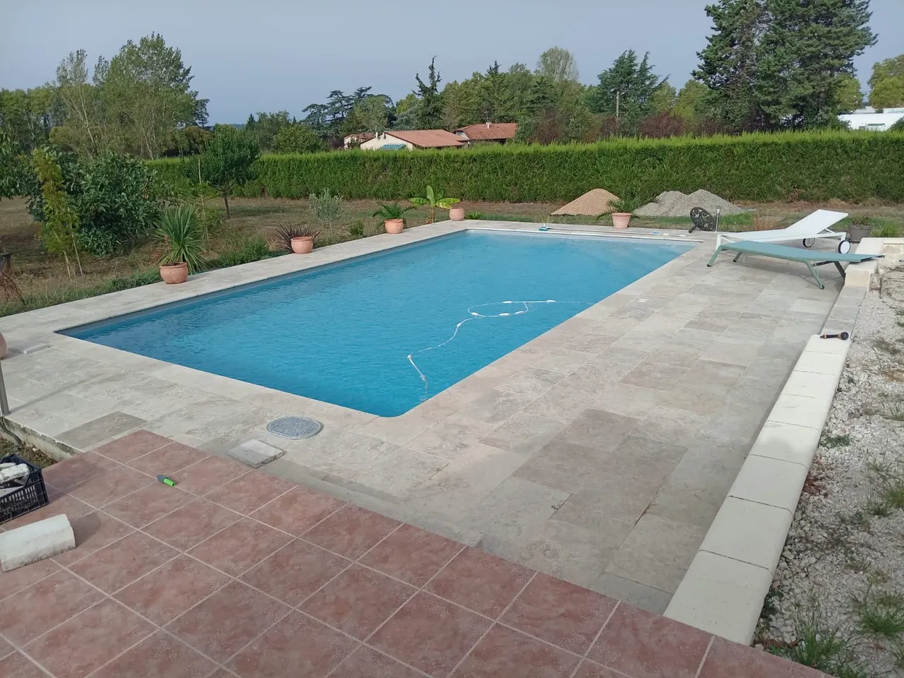 Piscine rectangulaire entourée d’une plage en dalles de pierre claire, terrasse en carreaux de terre cuite au premier plan, haie et transat au fond