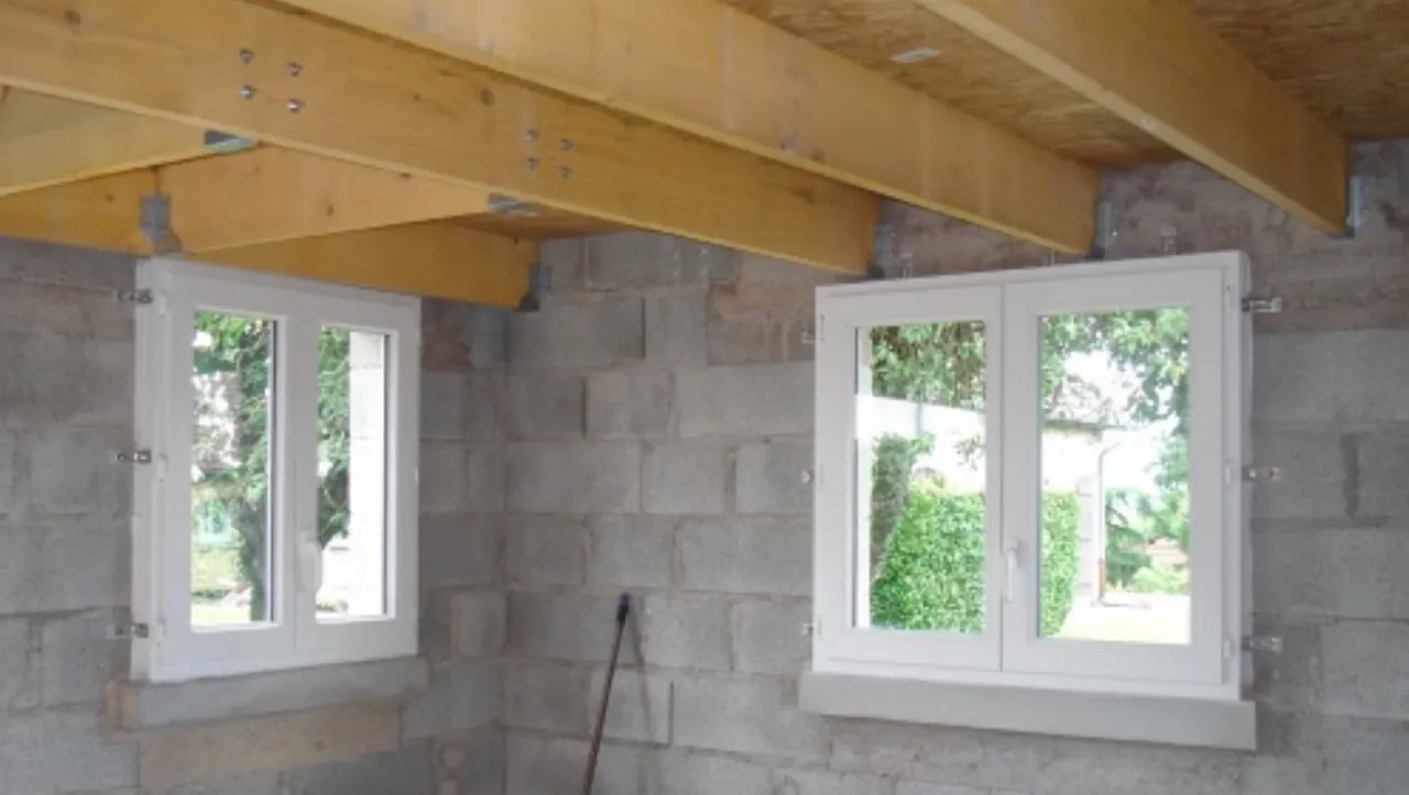 Intérieur d’un bâtiment en blocs béton : poutres en bois et plancher au-dessus, deux fenêtres blanches posées