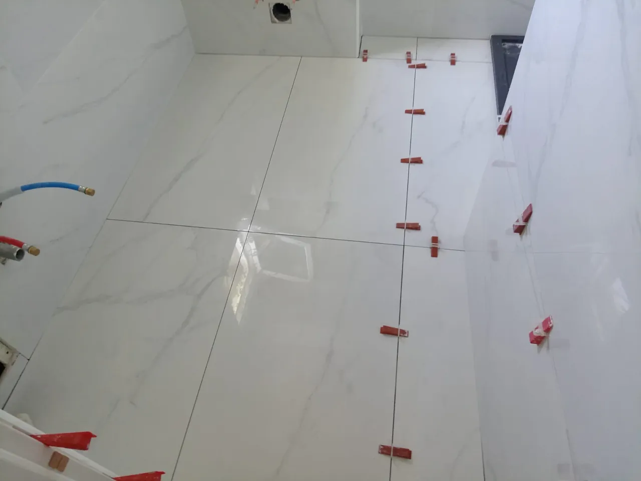 Salle de bains en cours de carrelage : grands carreaux blancs veinés au sol et aux murs, cales de nivellement rouges, attentes d’eau sur le mur