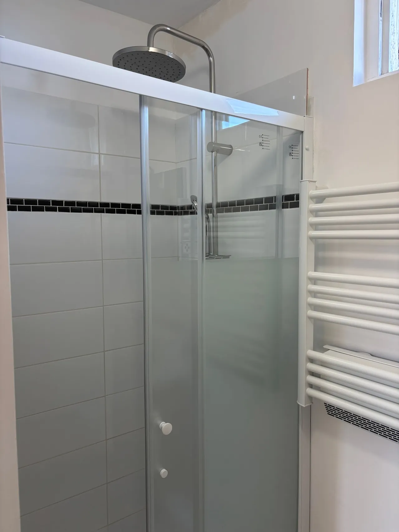 Douche terminée : porte coulissante vitrée, colonne de douche à pommeau, faïence blanche à frise noire, sèche-serviettes