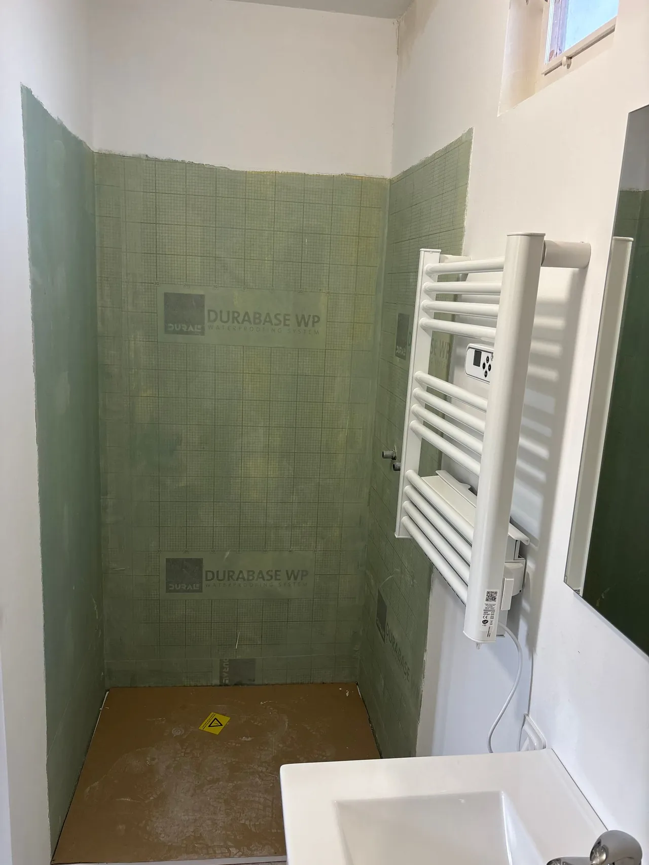 Salle d’eau en travaux : murs de la douche couverts de panneaux d’étanchéité verts, sèche-serviettes posé, vasque au premier plan