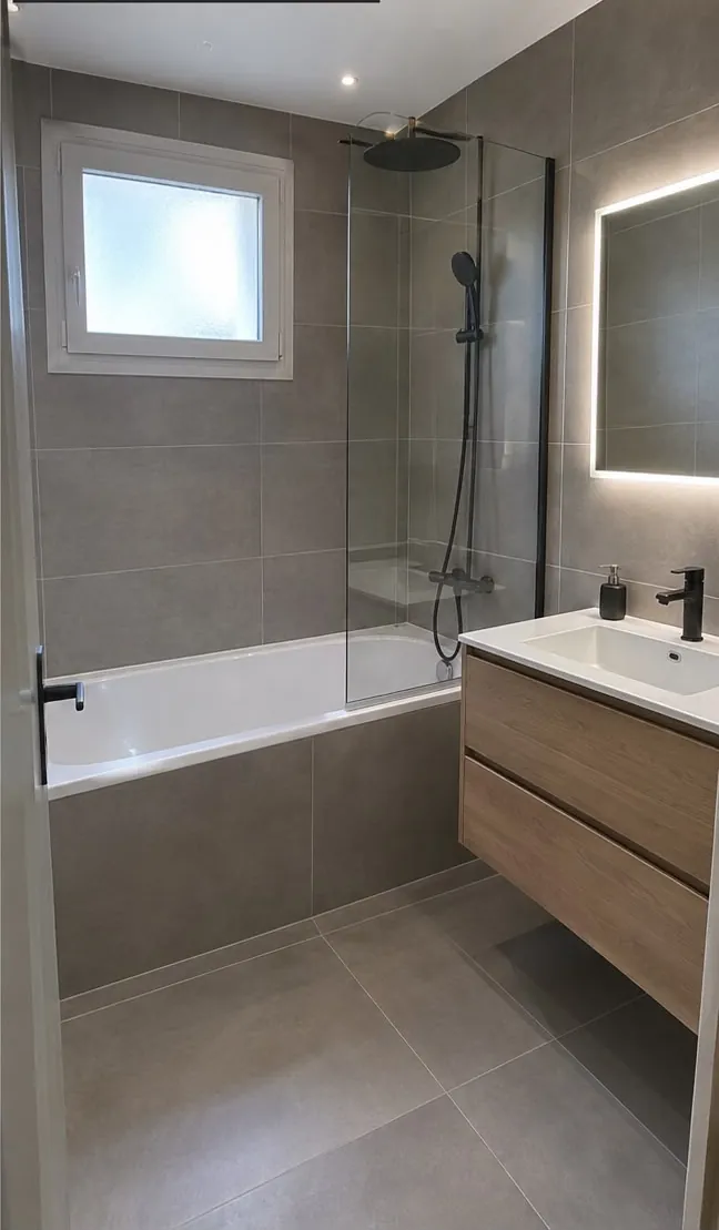 La même salle de bains refaite : grands carreaux gris, baignoire neuve et sa paroi vitrée, meuble vasque en bois, miroir lumineux
