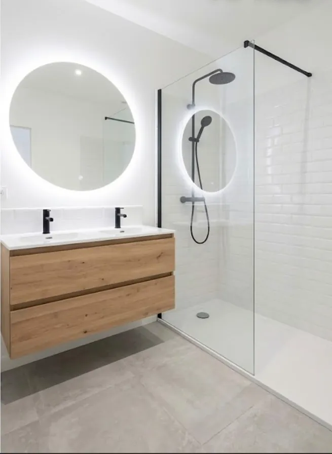 La même salle de bains refaite : douche à paroi vitrée, double vasque sur meuble en bois, miroir rond lumineux