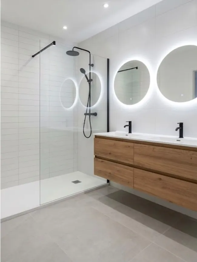 La même salle de bains refaite : douche à l’italienne vitrée, double vasque sur meuble en bois, deux miroirs ronds lumineux
