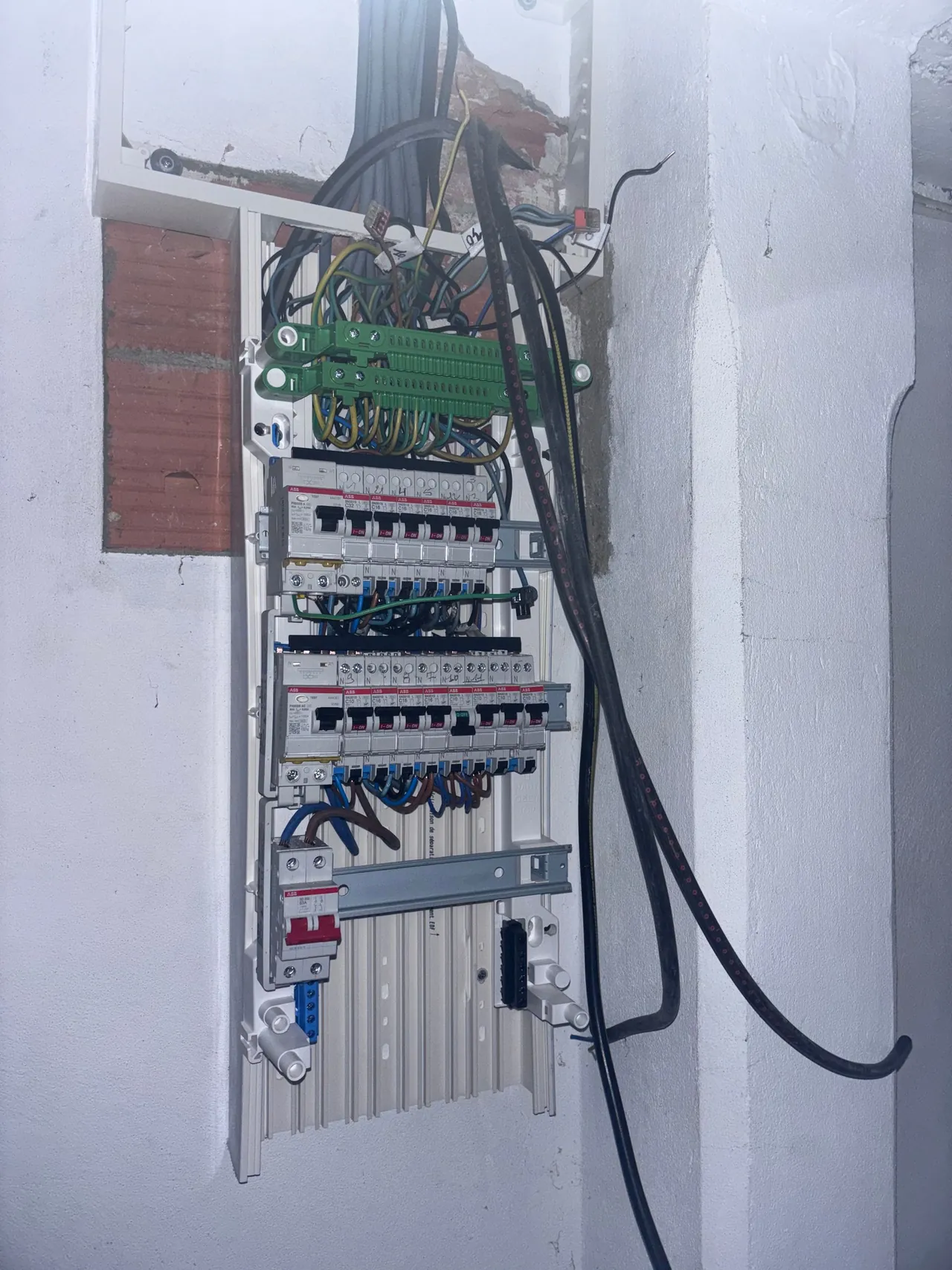 Tableau électrique en cours de câblage dans une niche en briques : rangées de disjoncteurs, peignes, borniers verts et câbles d’arrivée