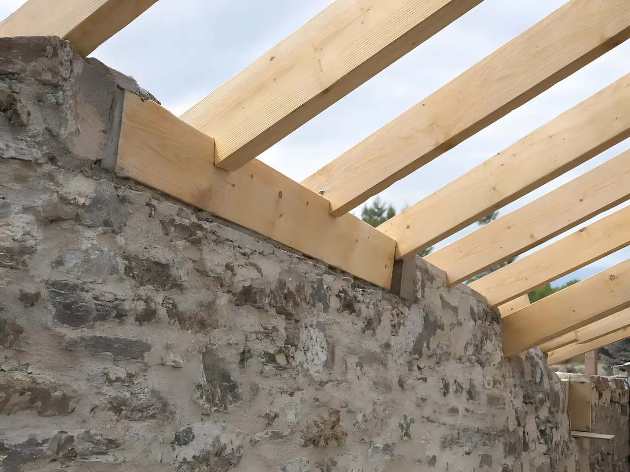 Panne neuve en bois clair calée en haut d’un vieux mur de pierre, chevrons posés dessus à intervalles réguliers