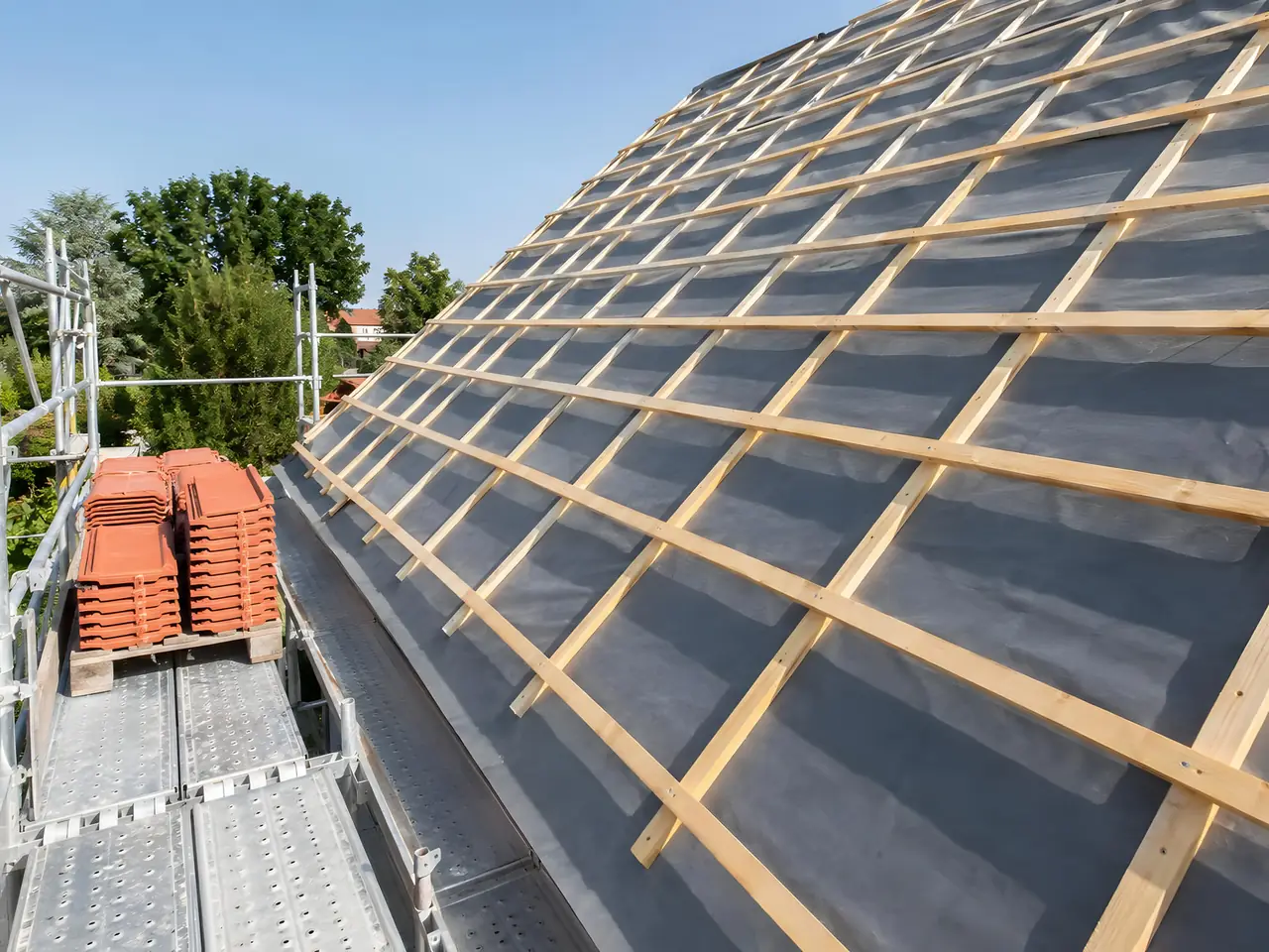 Pan de toit couvert d’écran sous-toiture gris et de liteaux neufs, piles de tuiles rouges posées sur l’échafaudage
