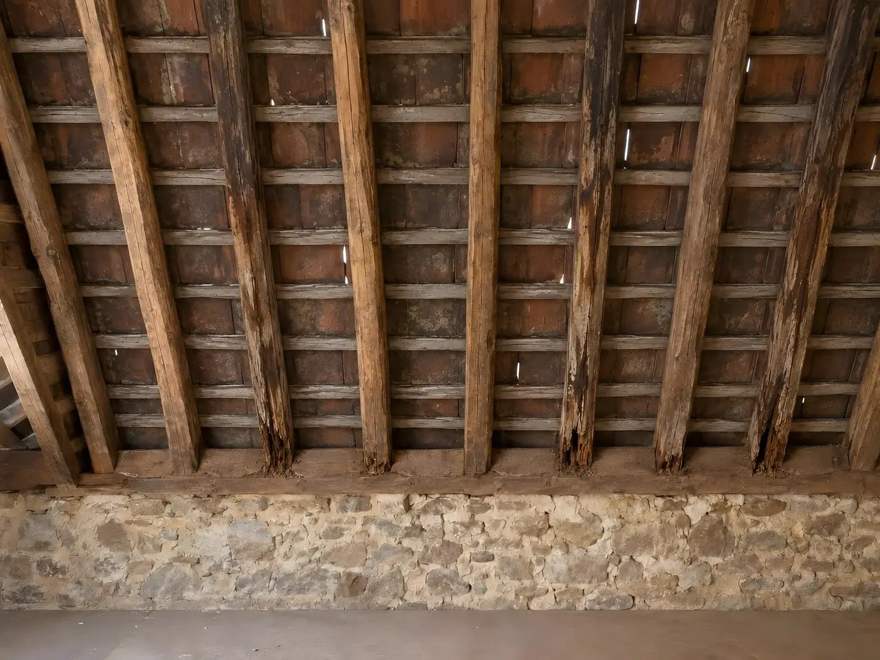 Charpente ancienne vue du grenier, chevrons usés et vieux liteaux sous des tuiles où passent des filets de jour