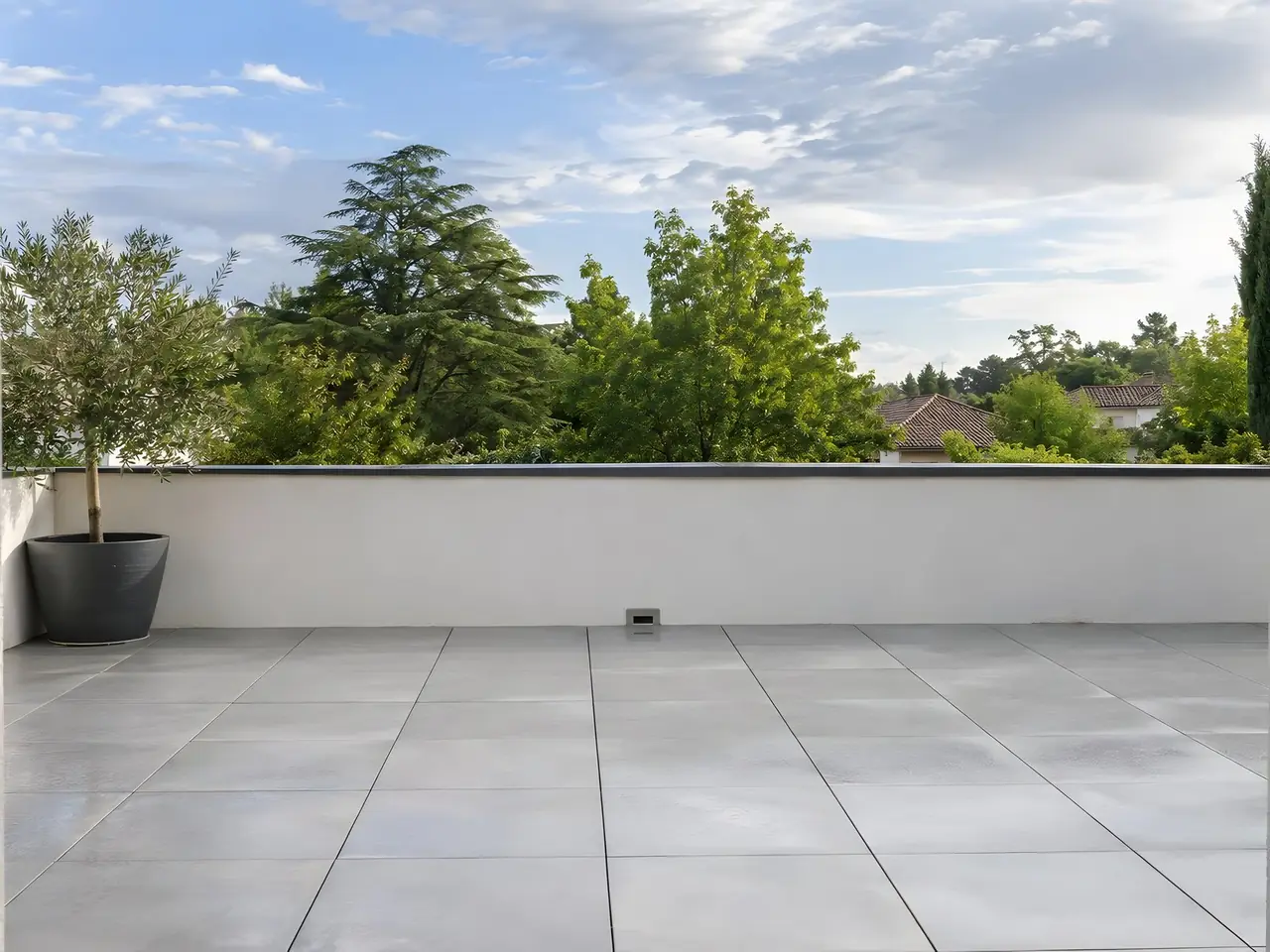 Terrasse en dalles gris clair sèches et régulières, muret enduit blanc, olivier en pot, cime d’arbres au-delà