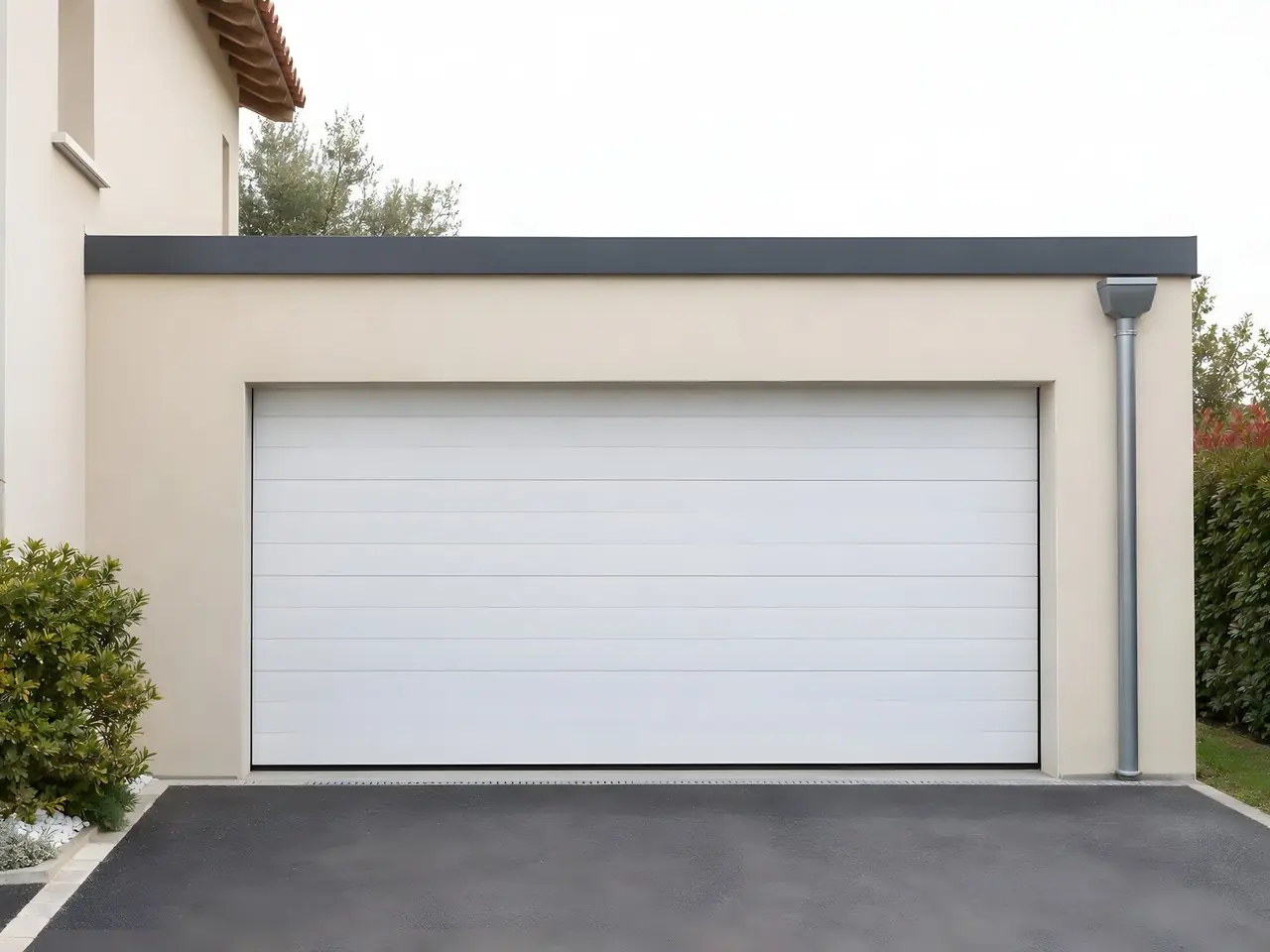 Garage à toit plat fini par une couvertine gris anthracite droite, enduit beige propre, porte sectionnelle blanche