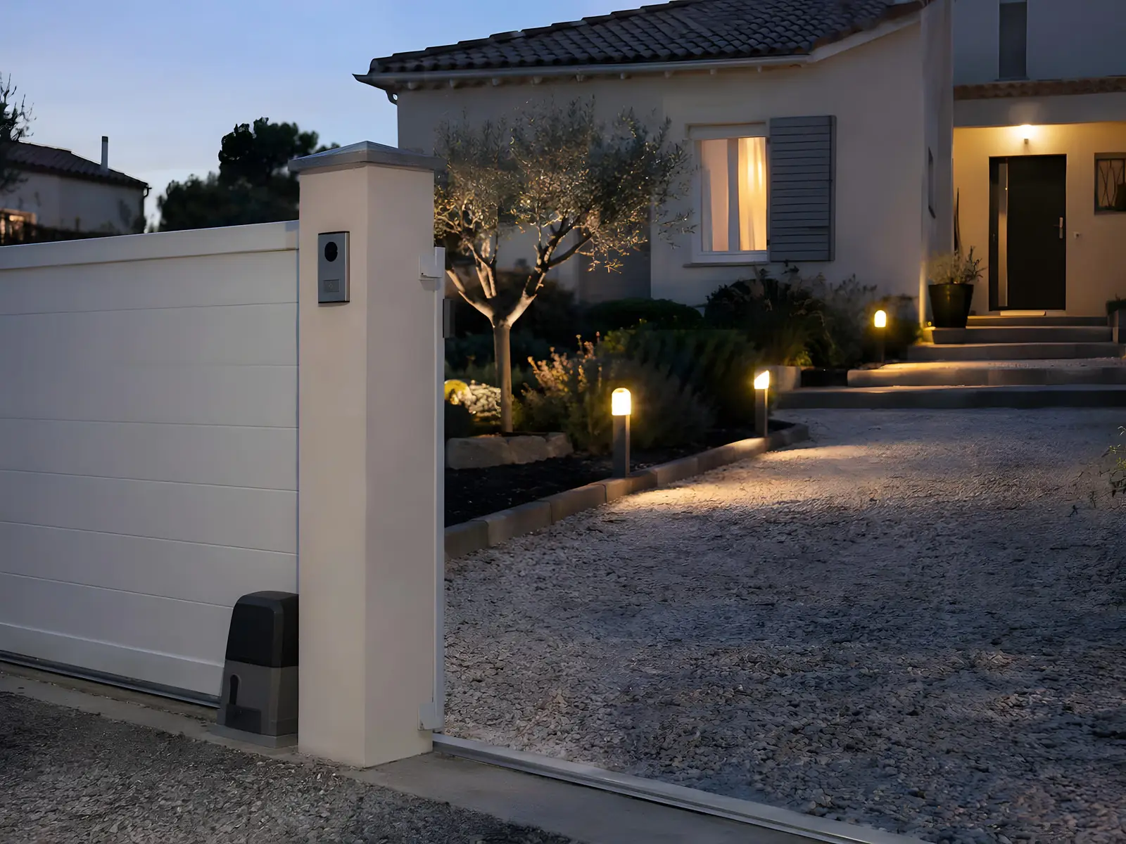 Portail coulissant blanc motorisé à la tombée du jour, visiophone sur le pilier et bornes lumineuses le long de l’allée