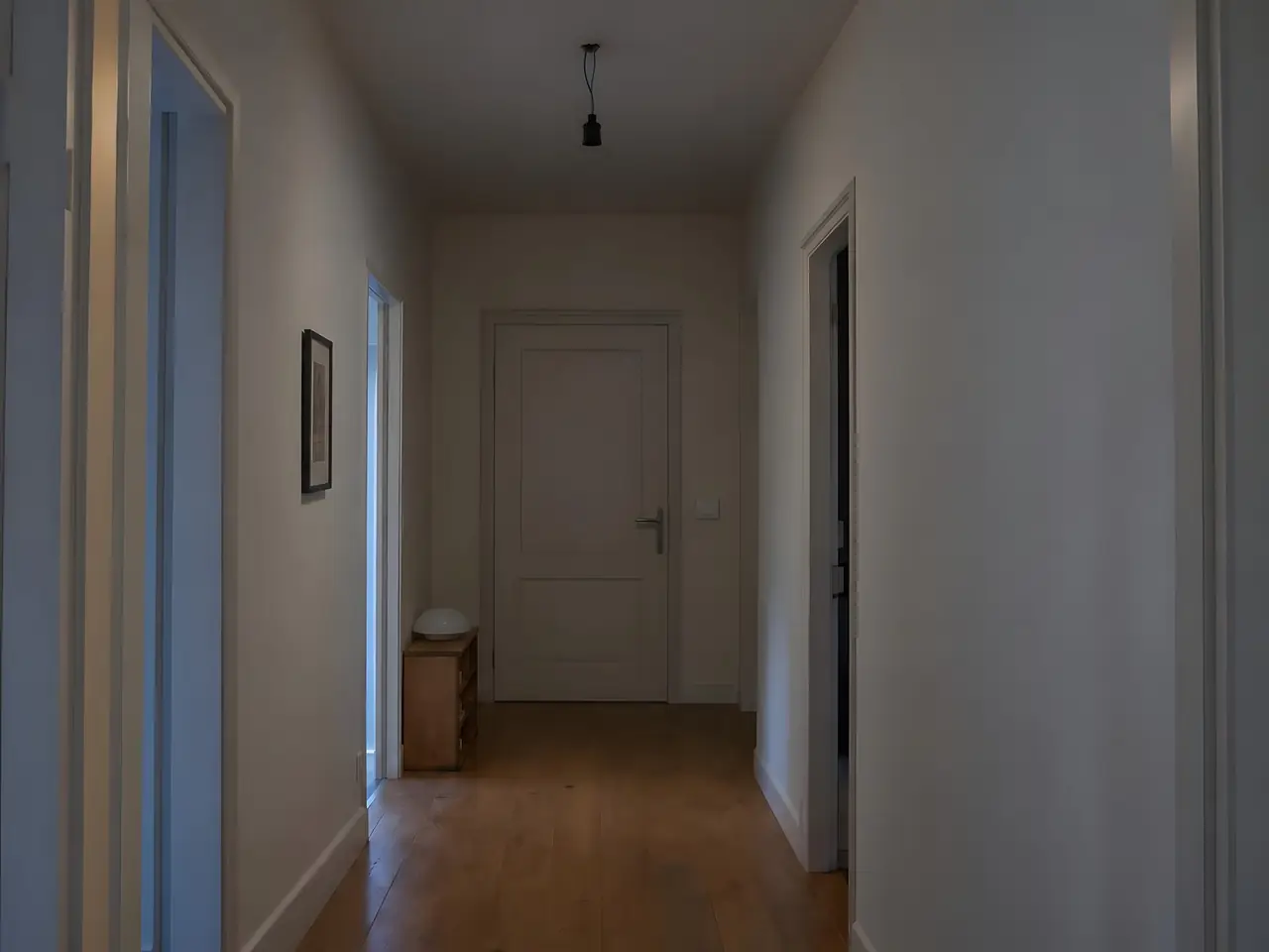 Couloir dans la pénombre, douille nue pendue au plafond par ses fils, verre opale posé sur le meuble à chaussures