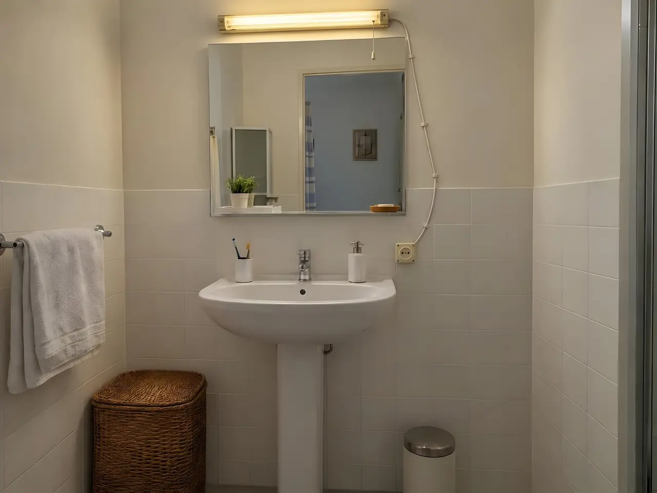 Lavabo sur colonne sous un miroir, vieille réglette jaunie, fil blanc apparent qui descend jusqu’à une prise en saillie
