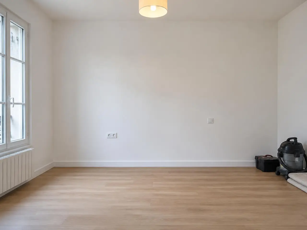 La même pièce aux murs blancs : prise et interrupteur posés, luminaire allumé, aspirateur rangé dans un coin