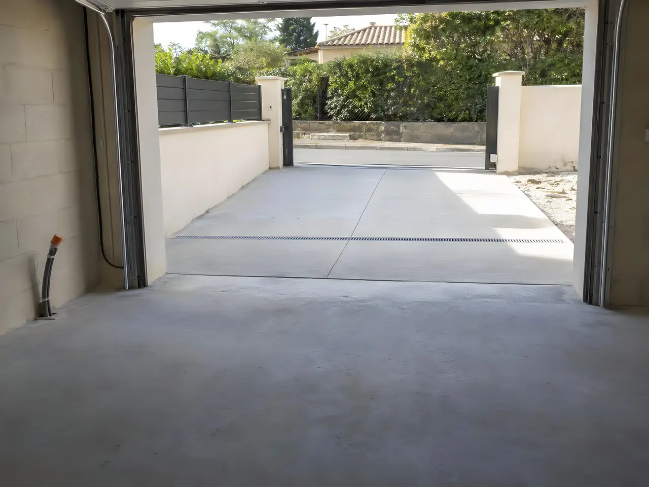 Intérieur d’un garage neuf vide : sol en béton, grande porte ouverte sur une allée bétonnée et un portail