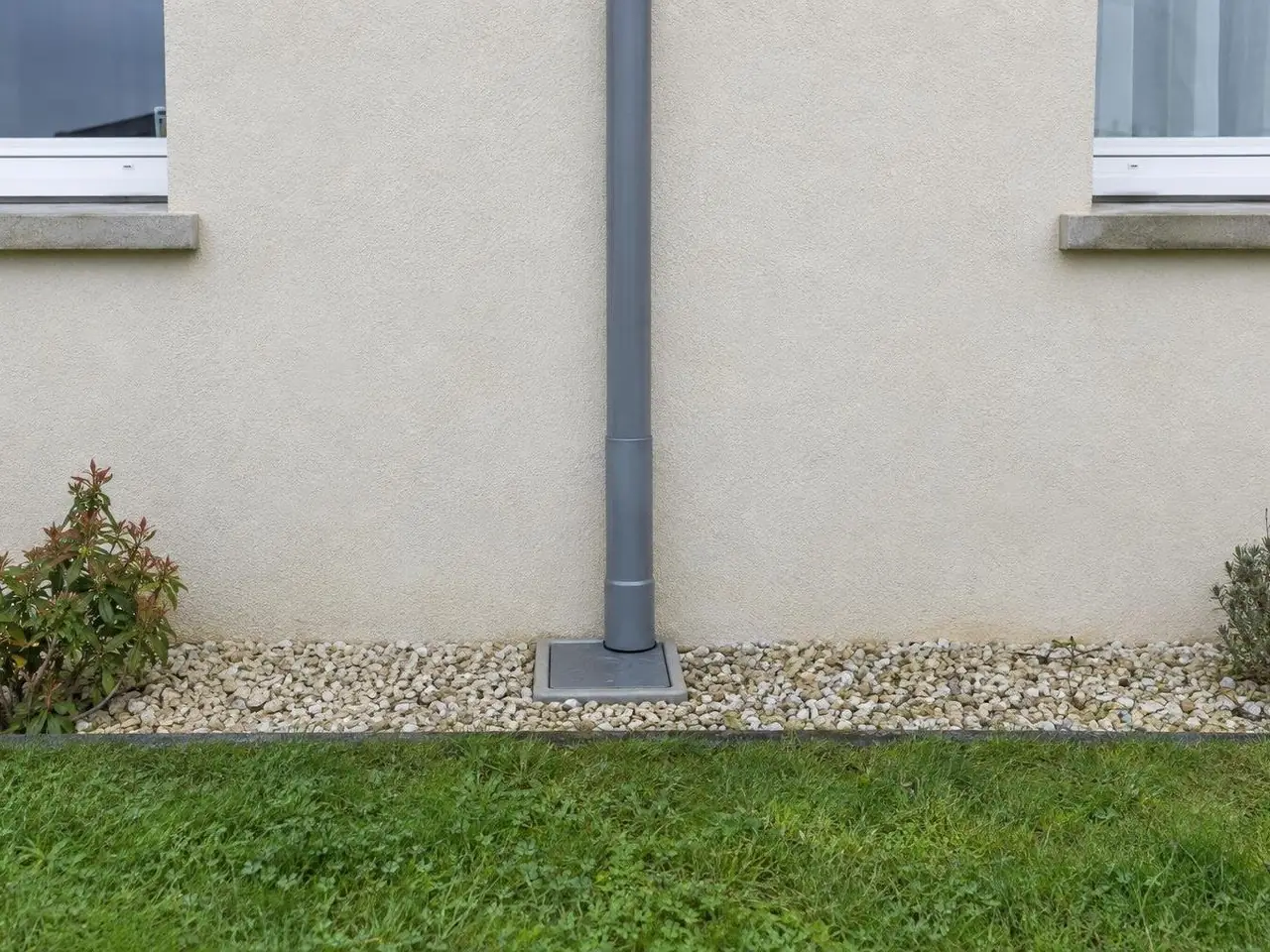Descente de gouttière grise raccordée à un regard carré au ras du sol, bande de graviers clairs au pied de la façade